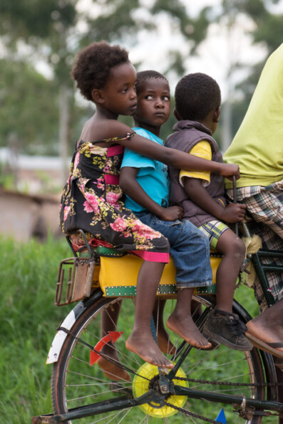 African kids pester each other while siting on the back rack of a bicycle taxi.