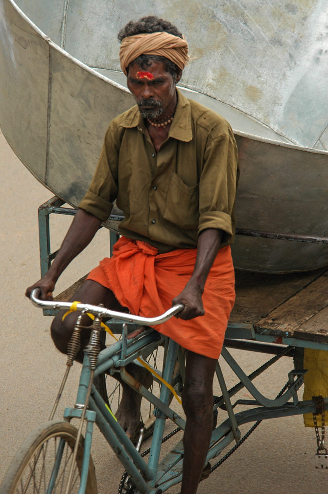 An Indian carries a large metal pot on the back of his rickshaw