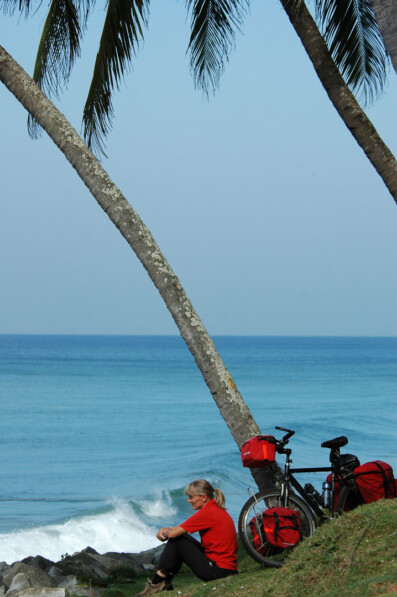 A Western cyclist sits looking out over the sea with their touring bicycle leaned against a palm tree in India.