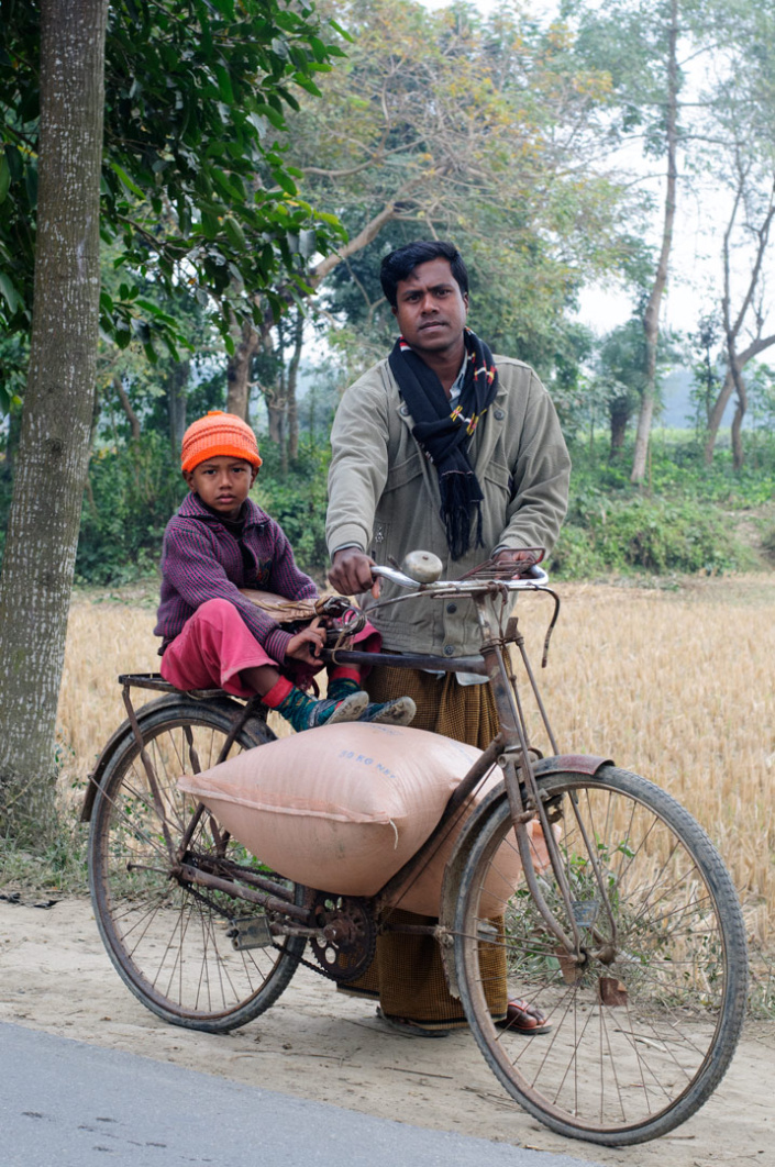 A father stands behind his bicycle while his son sits on the back rack
