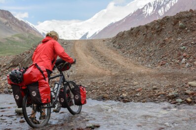 A red touring cyclist pushes their fully loaded bike through a creek near the border of Kyrgyzstan.