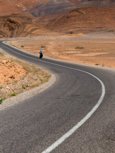 A small blue cyclist heads down a windy road in the Moroccan desert.