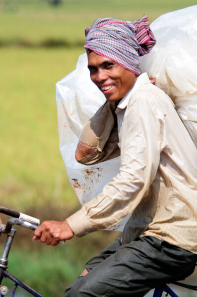 A Cambodian man cycles carrying a full bag on his shoulders.