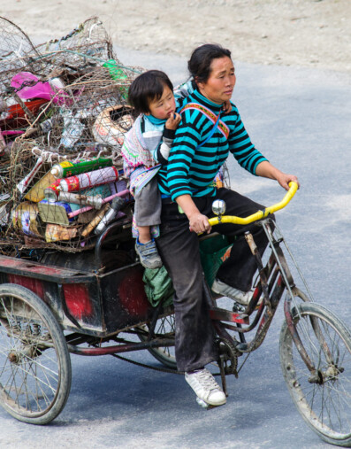 A Chinese lady pedals her cargo bike with her child attached to her bike.