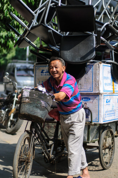 A Chinese man pushes his cargo bike that is loaded up with chairs.