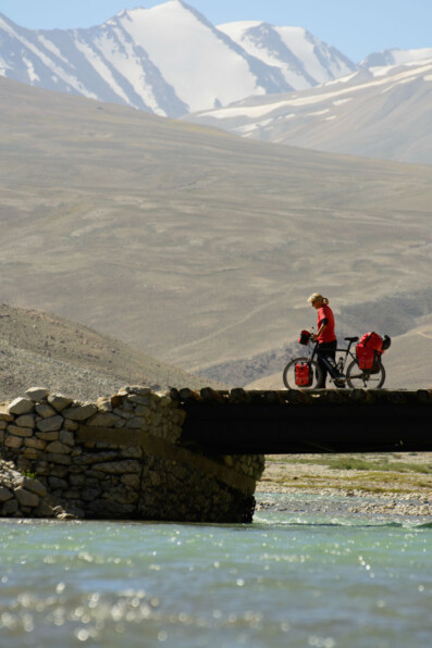 A touring cyclist stands on a bridge next to the Pamir highway.