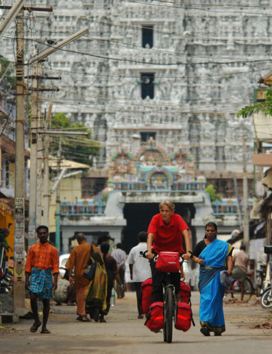 A western cyclist bicycles through an Indian street. In the background a South Indian temple – gopuram can be seen.