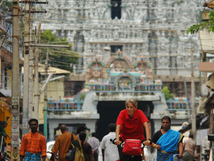 A western cyclist bicycles through an Indian street. In the background a South Indian temple – gopuram can be seen.