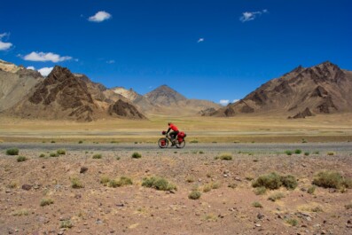 A red cyclist rides the Pamir highway with multicolored mountains behind them.