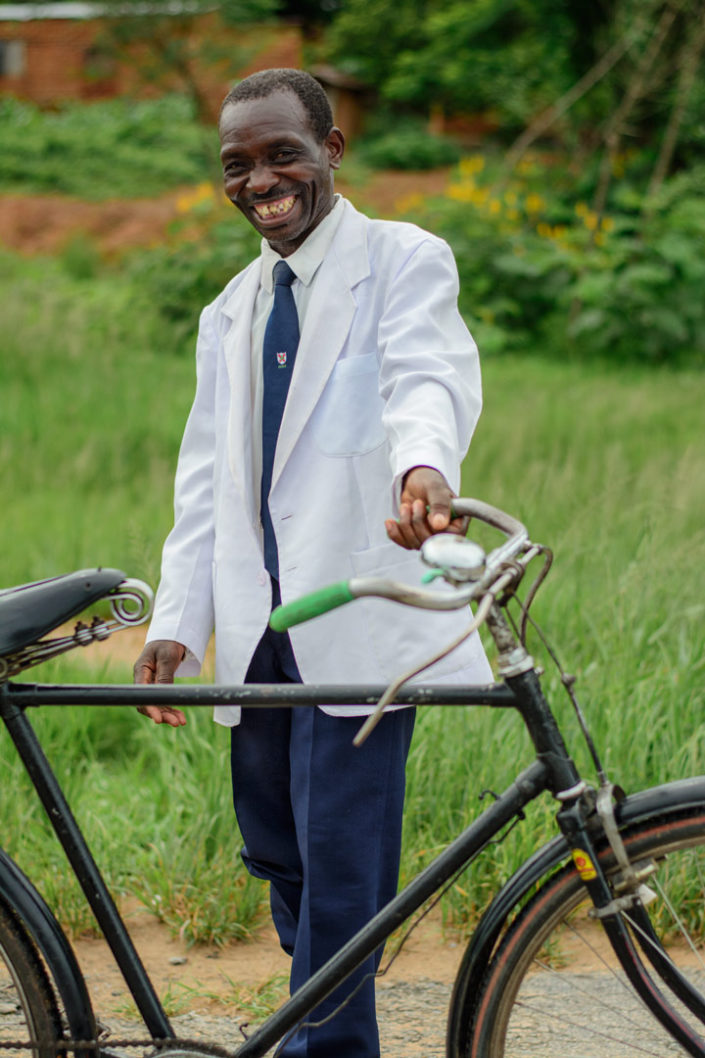 A respectably dressed man stands in front of a bicycle in africa.