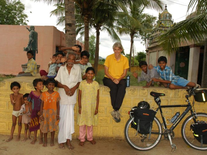 A western cyclist sits on a wall in South India and locals stand next to her.