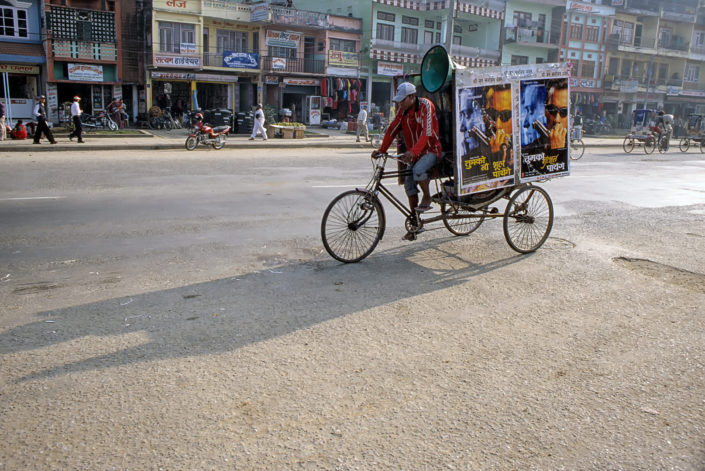 A chauffeur pedals his rickshaw covered with film posters through a town.