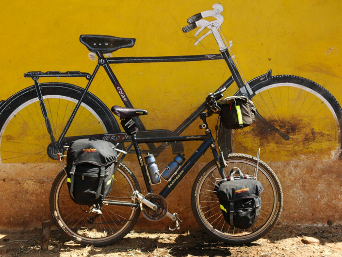 A touring bike is parked against a wall with a mural of an Indian Hero bicycle on it.