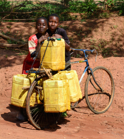 Two African children stand next to a bicycle loaded with water jugs.