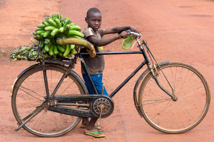 A young African boy stands next to his bike that has green bananas loaded on the back rack