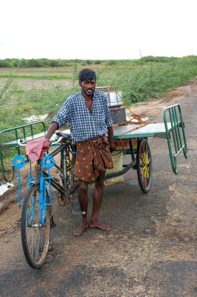 A man stands next to a rickshaw that is loaded up with a metal bed frame in India