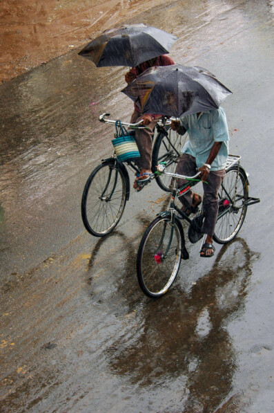 Two Indian cyclists pedal while holding umbrellas during monsoon rains